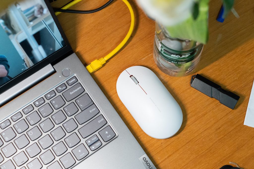 Hvid trådløs mus med scrollehjul, ved siden af en sølv Lenovo bærbar computer; på et lyst træbord.